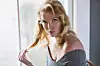 Seductive young blonde woman sitting indoors by sunny window, staring at the camera, playing with her hair.