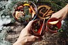 first person view of mans and womans hands holding glasses with Christmas hot mulled red wine with spices and fruits on a wooden rustic table background with spruce snow-covered branches. Traditional