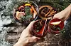 first person view of mans and womans hands holding glasses with Christmas hot mulled red wine with spices and fruits on a wooden rustic table background with spruce snow-covered branches. Traditional