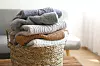 Bunch of stacked knitted pastel color sweaters with different knitting patterns perfectly folded in wicker basket on table, living room background. Fall winter season knitwear. Close up, copy space.