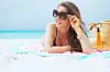 smiling stylish 40 year old woman in sunglasses and with long curly hair in elegant black bathing suit on a white beach looking into the distance and sun tanning.