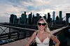 Žena u bijelom topu i sunčanim naočalama pozira na mostu ispred horizonta Manhattana.