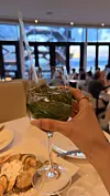 Ruka drži čašu zelenog koktela u restoranu uz more, uz pladanj morskih delicija na stolu.