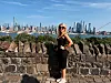 Žena u crnoj odjeći i sunčanim naočalama pozira ispred gradske panorame uz vodu.