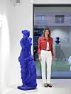 Žena u crvenom kariranom sakou, bijelim hlačama i crnim cipelama stoji uz plavu skulpturu u galeriji.