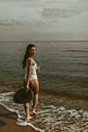 Žena u bijelom kupaćem kostimu i s crnim šeširom hoda uz morsku obalu.