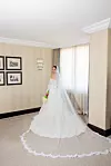 Mladenka u bijeloj čipkastoj vjenčanici i velu stoji uz prozor u elegantnoj sobi.