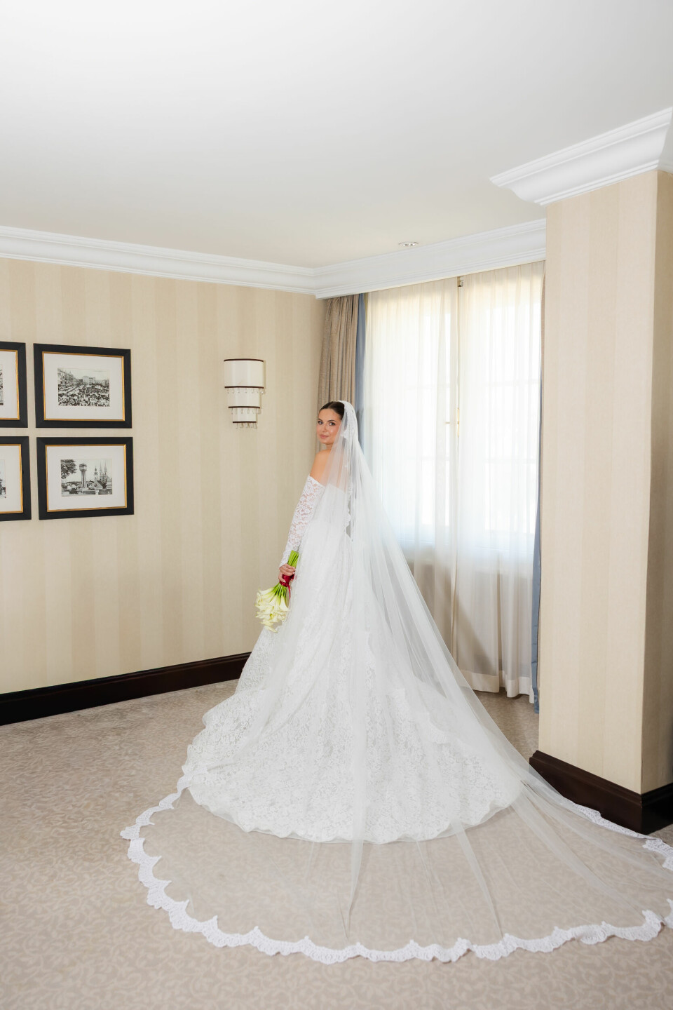 Mladenka u bijeloj čipkastoj vjenčanici i velu stoji uz prozor u elegantnoj sobi.