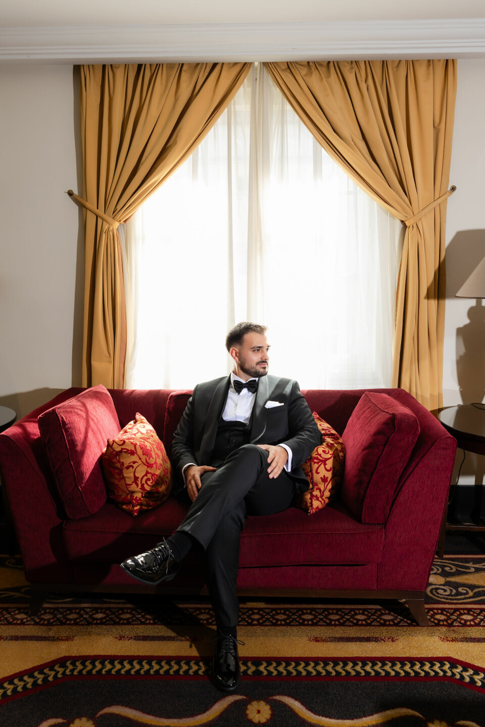 Muškarac u crnom smokingu sjedi prekriženih nogu na bordo sofi u elegantno uređenoj sobi.