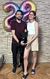 Muškarac i žena u ležernoj odeći poziraju s crnim psom ispred balona s brojem 28.