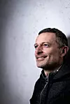 Juraj Šebalj u crnoj jakni pozira nasmiješen pred svijetlosivom pozadinom.