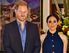 Princ Harry u tamnom odijelu i Meghan Markle u tamnoplavoj haljini poziraju u interijeru.