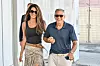 George i Amal Clooney hodaju uz zračnu luku; Amal nosi crni top i suknju, a George polo majicu i hlače.