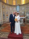 Mladenci u tamnoplavom odijelu i bijeloj vjenčanici poziraju pred oltarom.
