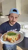 Brooklyn Peltz Beckham u plavoj kapi drži tanjur pilećeg parmezana u svijetloj kuhinji.
