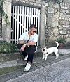 Muškarac u sunčanim naočalama sjedi uz kameni zid, a kraj njega stoji bijelo-smeđi pas.