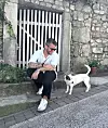 Muškarac u sunčanim naočalama sjedi uz kameni zid, a kraj njega stoji bijelo-smeđi pas.