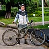 Muškarac u srebrno-crnoj jakni stoji uz crni brdski bicikl u zelenom parku.