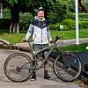 Muškarac u srebrno-crnoj jakni stoji uz crni brdski bicikl u zelenom parku.