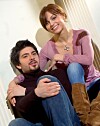 Nasmiješena žena u ružičastoj majici i muškarac u tamnoj majici poziraju sjedeći u interijeru.