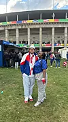 Dvoje navijača u sportskoj odjeći pozira na travnjaku ispred velikog stadiona.