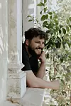 Nasmiješen muškarac u crnoj majici naslonjen na prozor među zelenim lišćem.