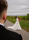 Mlada u bijeloj vjenčanici s crvenim buketom stoji na putu kroz vinograde, dok je mladoženja u prvom planu.