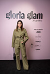 Žena u maslinastom odijelu pozira na događanju Gloria Glam uz ružičastu pozadinu i logotipe partnera.