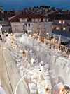 Svečano uređena terasa s dugim bijelim stolovima, cvijećem i rasvjetom iznad krovova povijesnog grada.