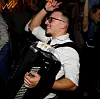 Nasmiješeni muškarac u bijeloj košulji svira crnu harmoniku među okupljenim ljudima.