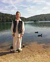 Mladić u bijeloj odjeći i crnom prsluku stoji uz jezero s patkama i šumovitim brdima u pozadini.