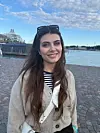 Mlada žena u bež kardiganu i prugastoj majici pozira uz riječnu luku.