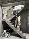 Osoba u crnoj kožnoj jakni i dugom smeđem kaputu pozira na betonskim stubama.