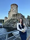 Žena u bijeloj bluzi i tamnoplavoj suknji stoji ispred kamene tvrđave uz vodu.