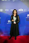 Žena u dugoj crnoj haljini pozira s trofejem na crvenom tepihu ispred brendiranog zida.