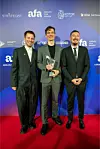 Troje muškaraca u odijelima pozira na crvenom tepihu; muškarac u sredini drži trofej.