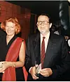 Žena u crveno-crnoj haljini i muškarac s naočalama u tamnom odijelu na događanju.