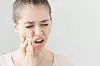 Close headshot of young European female isolated on grey background suffering from severe toothache, feeling pain so strong that she is pressing fingers to cheek to calm it down, looking desperate.